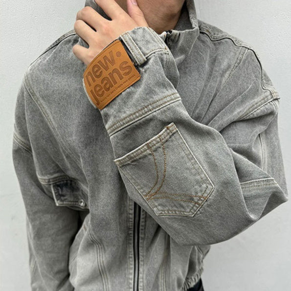 Person wearing a gray denim jacket with a brown patch on the sleeve, against a plain background.